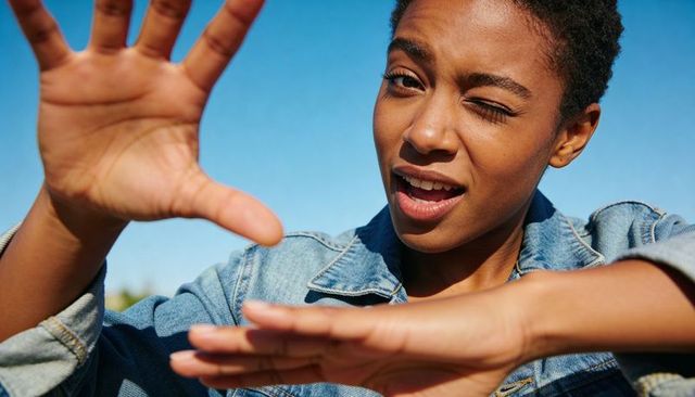 Winking woman playfully framing face with hands in sunlit denim jacket against blue sky