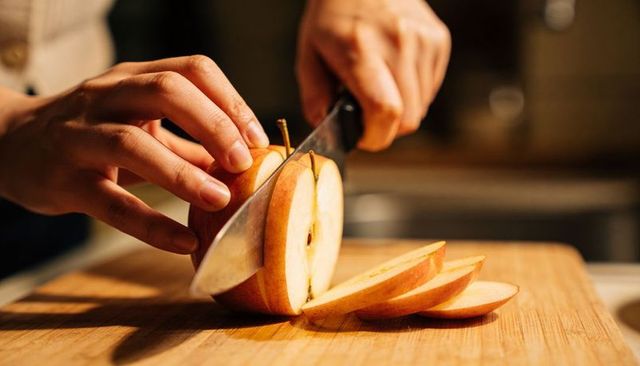 Hands slicing ripe pear on wooden cutting board with chef knife closeup, warm lighting
