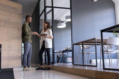 Diverse professionals shaking hands by glass wall holding tablet in modern workplace