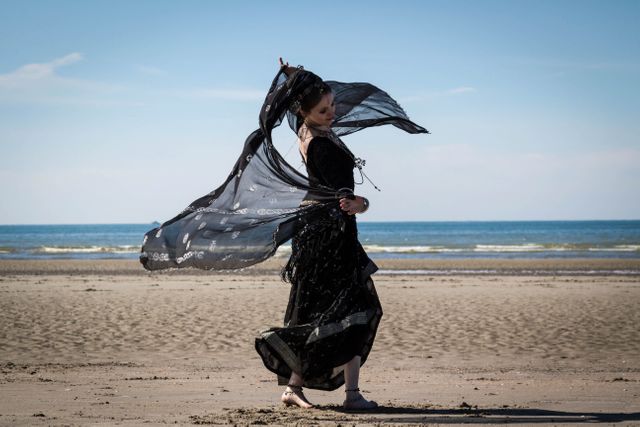 Woman in a black dress dancing on a sandy beach with a flowing scarf. The ocean waves crash in the background against a blue sky. Use for promoting summer fashion, vacations, travel destinations, or concepts of freedom and relaxation.