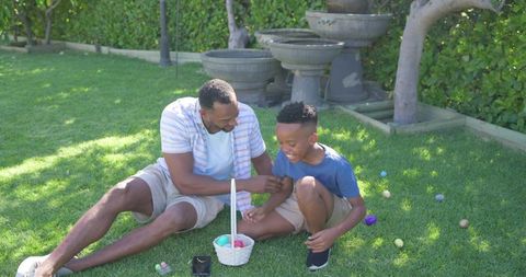 African american father and son playing with easter eggs on backyard lawn, candid moment