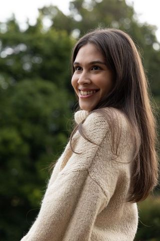 Smiling young woman wearing cream knit sweater looking over shoulder in leafy park portrait