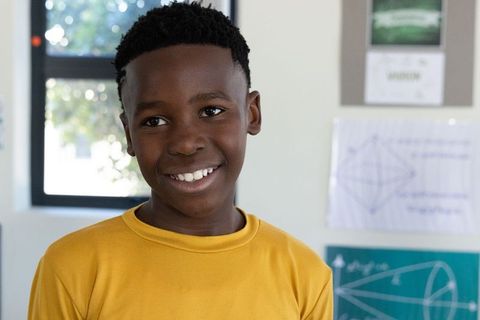 Smiling african american boy in math classroom wearing mustard t-shirt standing by window