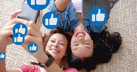 Two friends lying on floor taking selfies with floating social media like icons