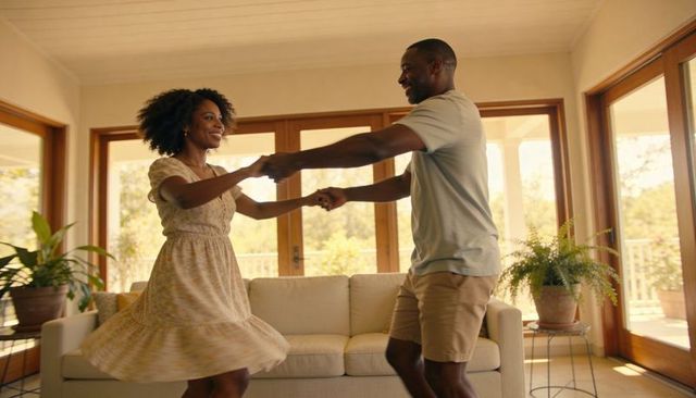 Sunlit couple dancing and spinning in cozy living room with sofa and indoor plants