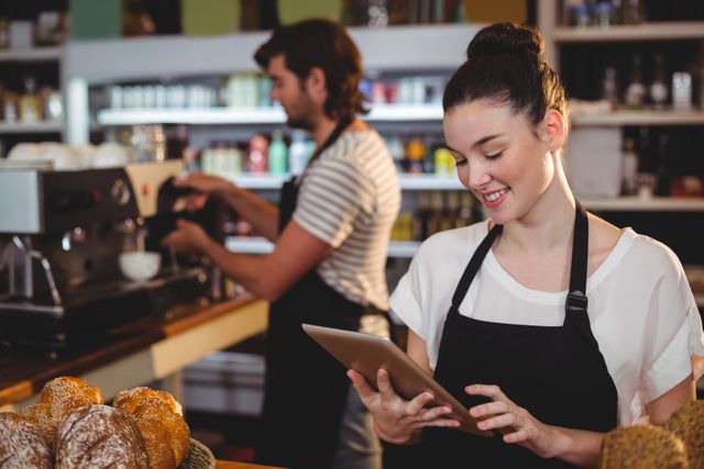Waitress in a cozy cafe smiling while using a digital tablet, with a barista working in the background. Ideal for marketing materials highlighting customer service, teamwork in small businesses, and the integration of technology in cafes and bakeries.
