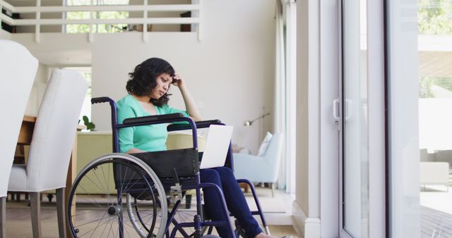 Thoughtful biracial disabled woman in wheelchair using laptop in living room. wellbeing and domestic lifestyle with physical disability.