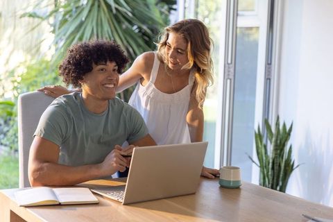 Sunlit remote work with smiling diverse couple collaborating on laptop at home desk