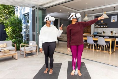 Diverse women practicing vr yoga on patio with open-plan kitchen and headsets for wellness lifestyle