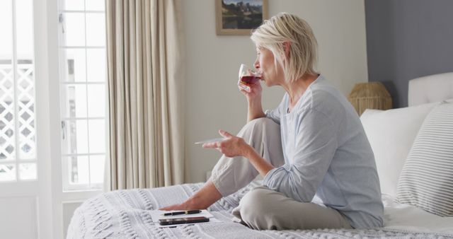 Image shows an older woman sitting on a bed at home, holding a cup of tea and relaxing. She is enjoying a quiet moment and looking away thoughtfully. This can be used for articles or promotions on senior lifestyle, health and wellness, morning routines, relaxation, and home decor.