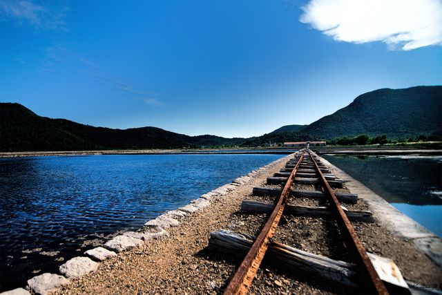Rustic railway tracks lead towards a small lakehouse, surrounded by serene waters reflecting the clear blue sky. Majestic mountains in the background complete this breathtaking landscape. Ideal for travel blogs, adventure tourism promotions, and nature-themed websites.