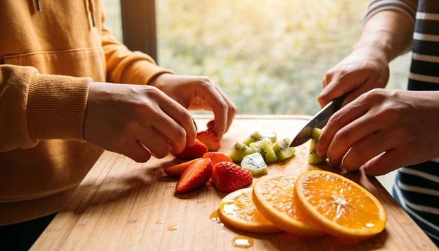 Sunlit kitchen hands slicing and arranging strawberries, kiwi and orange on wooden cutting board