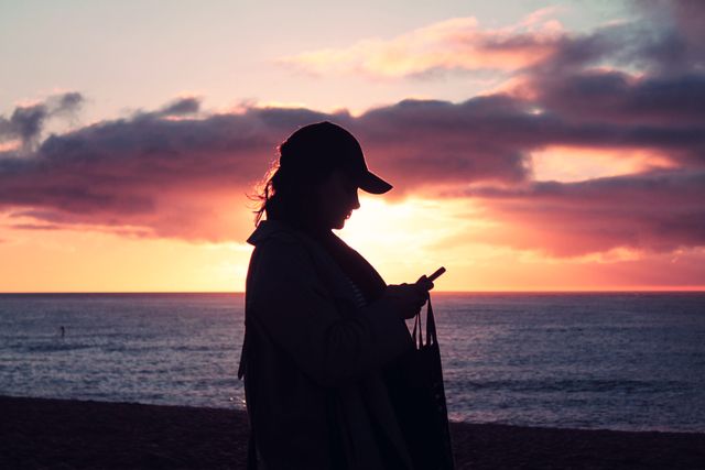 Silhouette of a woman looking at her smartphone against the backdrop of a breathtaking beach sunset. The sky is adorned with vibrant shades of pink and purple, contrasting beautifully with the calm sea. Ideal for concepts related to technology, relaxation, solitude, and the beauty of nature. Can be used for travel blogs, lifestyle websites, advertisements for beach resorts, and social media content focused on digital connectivity and nature.