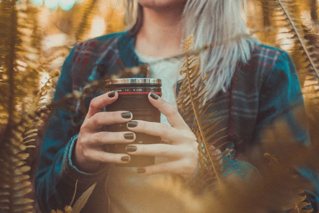 Person holding a mug designed like a camera lens while surrounded by autumn foliage. They are wearing a plaid jacket. Captures a cozy, outdoorsy feel. Excellent for use in blogs about travel, nature photography, cozy fall moments, and lifestyle photography.
