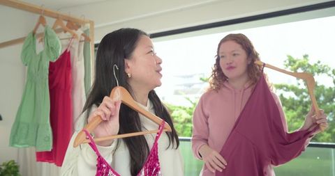 Diverse friends comparing pink sequin and maroon dresses in bright balcony closet