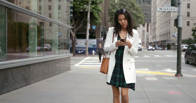 Young Asian woman checks her phone on a city street, with copy space. She's dressed smartly, suggesting she might be on a break from work or school.