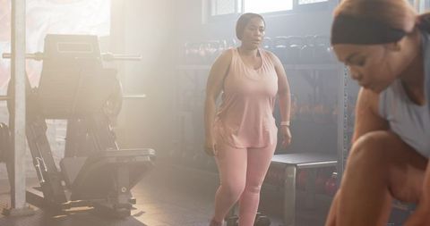 Backlit diverse mid-adult women working out, adjusting shoes near bench and kettlebells