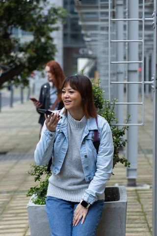 Asian woman talking into smartphone while sitting on concrete planter in urban plaza