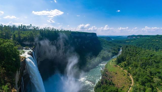 Panoramic cascading waterfall plunging from cliff into misty gorge with winding river and forest tra