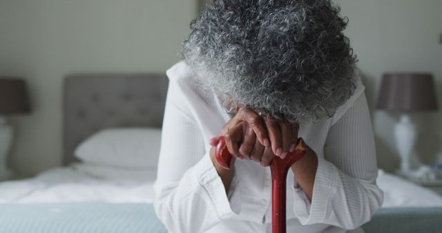 Elderly woman is grieving while sitting on a bed and resting her head on a cane. Her overall demeanor suggests deep sadness and loneliness. This image can be used to portray themes of grief, depression, old age, and the emotional struggles often faced by seniors. It is suitable for articles and campaigns about elderly care, mental health awareness, and support services for the older population.