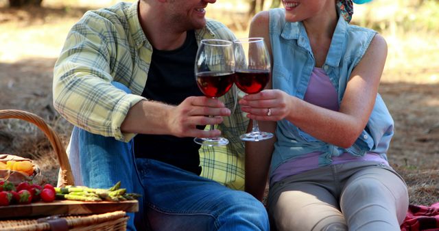 A young Caucasian couple enjoys a romantic picnic, toasting with glasses of red wine, with copy space. Their casual outdoor setting suggests a relaxed and intimate moment shared between the two.