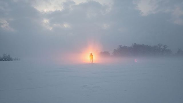Solitary figure standing on snowfield backlit by warm halo in foggy winter landscape