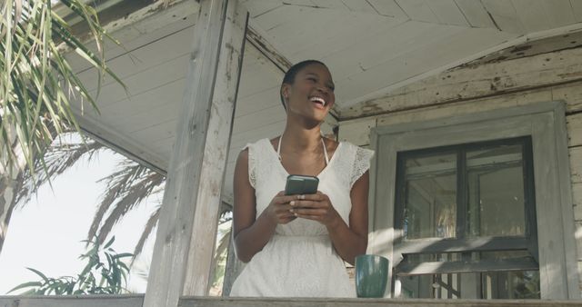 An image depicting a happy woman standing on a rustic porch, holding a smartphone and smiling brightly. She appears relaxed and enjoying the moment in a tropical, casual setting. Ideal for use in advertisements, lifestyle blogs, online content about happiness, mobile technology, summer activities, or relaxed home living.