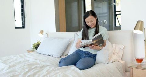 Asian woman reading on bed in minimalist sunlit bedroom, sipping tea, relaxing at home