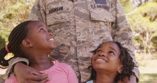 Father wearing Air Force uniform embraces daughters in outdoor setting. This depiction of family bond and military life is ideal for use in emphasizing togetherness, military family lifestyle, parental love, and outdoor joy within inclusive African American contexts. Great for use in promoting family-centered military services, educational materials, and celebratory events such as Veterans Day or Father’s Day.