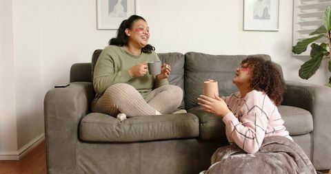 Two women laughing on gray sofa sharing coffee and enjoying cozy home conversation