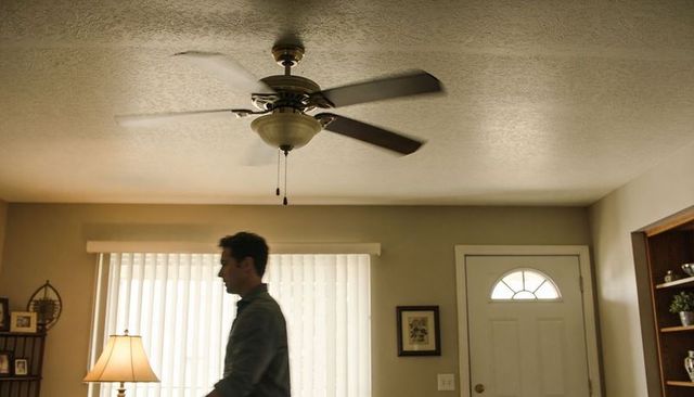 Walking man crossing living room with ceiling fan, vertical blinds and entryway silhouette