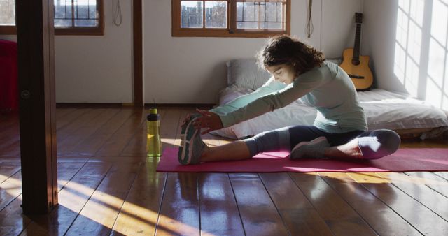 Young woman stretching on a pink yoga mat in a bright room with wooden floors. This setting indicates a home workout routine focused on flexibility and well-being. The presence of a water bottle suggests hydration during the physical activity. Natural sunlight streaming through windows enhances the calm and serene atmosphere, making this image suitable for articles about fitness at home, wellness blogs, and health-related advertisement campaigns.