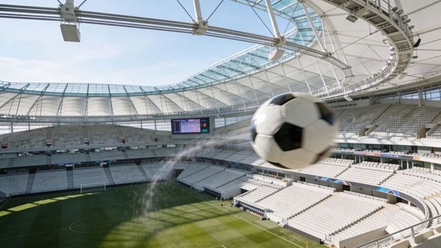 Wet soccer ball flying toward camera with water spray, motion blur in sunlit stadium