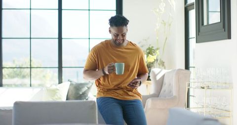 African american man sipping coffee and checking phone in sunlit modern living room