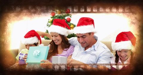 Happy family unwrapping christmas gifts on cozy sofa by decorated tree wearing santa hats