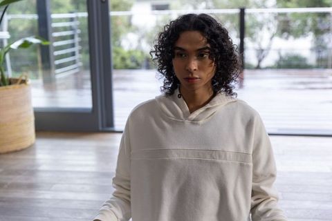 Non-binary person sitting on floor near sliding glass doors in cream hoodie practicing mindfulness