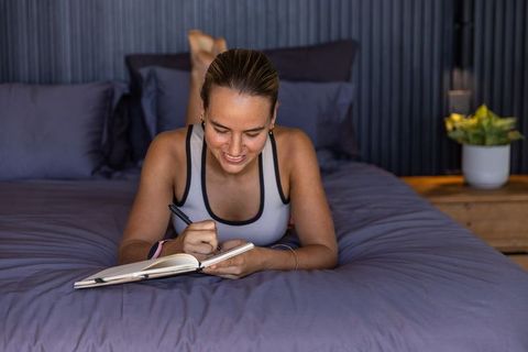 Young woman journaling on bed in sports bra relaxing with smartwatch in cozy modern bedroom