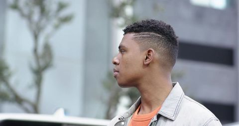 Young man standing on city sidewalk looking left wearing denim jacket and orange sweater