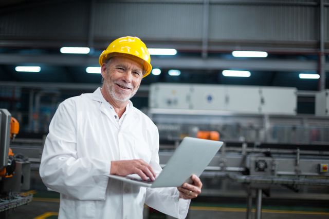 Senior factory engineer wearing safety helmet and white coat using laptop in a production plant. Ideal for illustrating industrial technology, manufacturing processes, quality control, and engineering roles in factories. Useful for articles, advertisements, and presentations related to industrial work, factory management, and technological advancements in manufacturing.