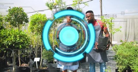 Two Nursery Workers Consulting Notepad While Holding Sapling in Black Pot in Greenhouse