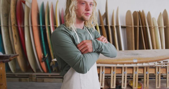 Thoughtful caucasian male surfboard maker with long blonde hair in apron at workshop, unaltered. Small business, work, sports equipment and craftsmanship.