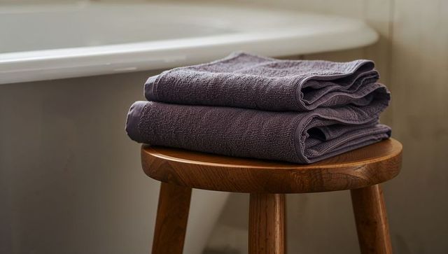 Folded plum towels on wooden stool beside bathtub minimalist bathroom linen texture