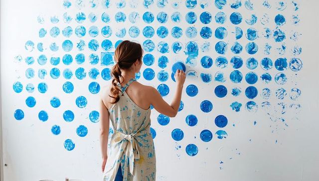 Female artist creating large blue circle mural using round stamps on white studio wall