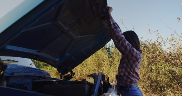 A woman is raising the hood of a car to check the engine outdoors in a rural area under sunny skies. She wears a plaid shirt and blue jeans, suggesting an adventurous spirit or dealing with a car breakdown. The image highlights the themes of independence, understanding mechanical issues, and being prepared during travel. This photo can be used in articles about car maintenance, empowerment, rural lifestyle, or travel blogs focusing on adventure and preparedness.