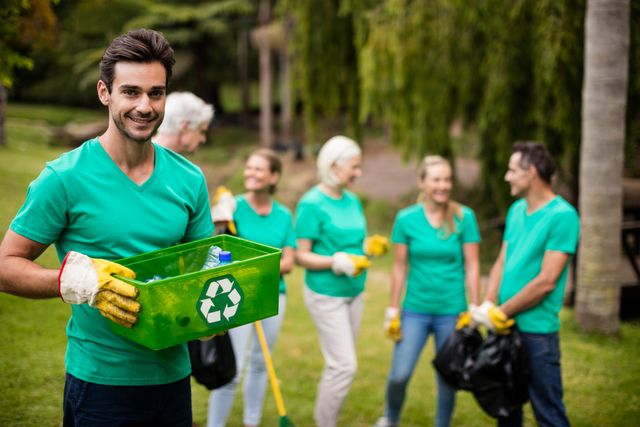 Smiling volunteer holding recycling bin in park with team in background. Ideal for promoting environmental awareness, community cleanup events, and sustainability initiatives.
