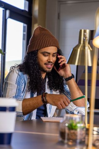 Young man in beanie talking on phone while pointing pencil at notebook in home office
