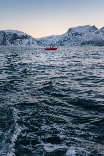 Dramatic photograph of a solitary red boat on icy waters with snow-capped mountains in the background, capturing the essence of winter isolation and tranquil scenery. Ideal for use in travel brochures, winter-themed backgrounds, or articles about remote and scenic nature destinations.