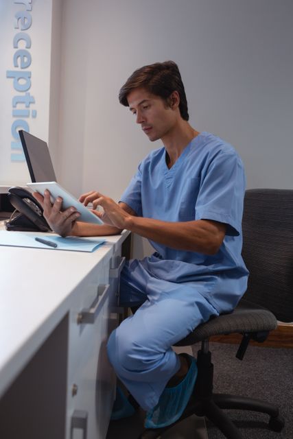 Caucasian male surgeon in blue scrubs using a digital tablet while seated at a desk in a hospital. Ideal for use in articles or advertisements related to healthcare technology, modern medical practices, hospital administration, and professional medical environments.