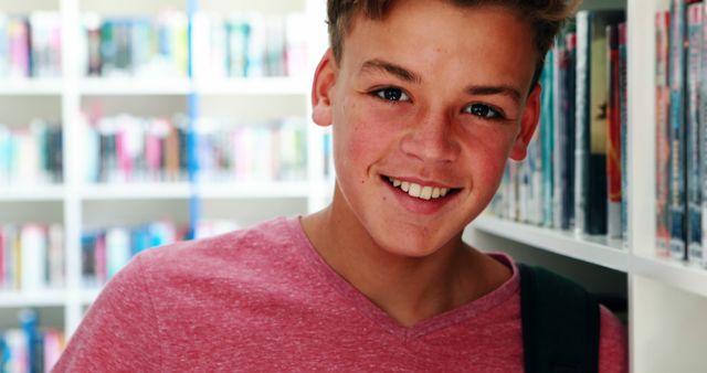 Teenage boy in a library smiling next to bookshelves, wearing a casual pink shirt. Ideal for educational materials, school brochures, and youth programs. Emphasizes positivity and learning in academic settings.