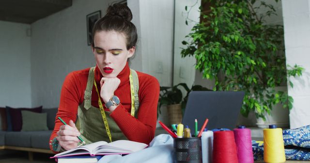 Fashion designer drawing sketches in notebook while sitting at desk with sewing supplies. Surrounded by vibrant threads and a green plant, indicating a creative and organized workspace. Suitable for use in articles about home studios, fashion design, creative workspaces, or maker communities.